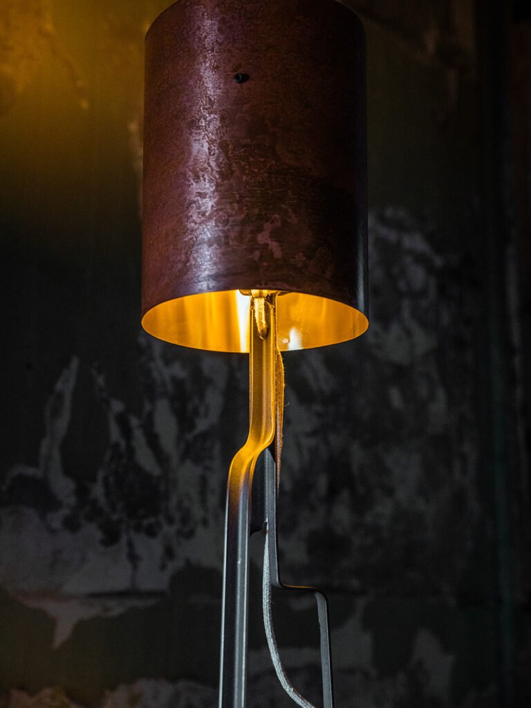 Close-up of a cylindrical brass lampshade with a textured, rose-copper patina and gold interior, emitting a warm yellow light. Background features a rough, dark wall with peeling paint.