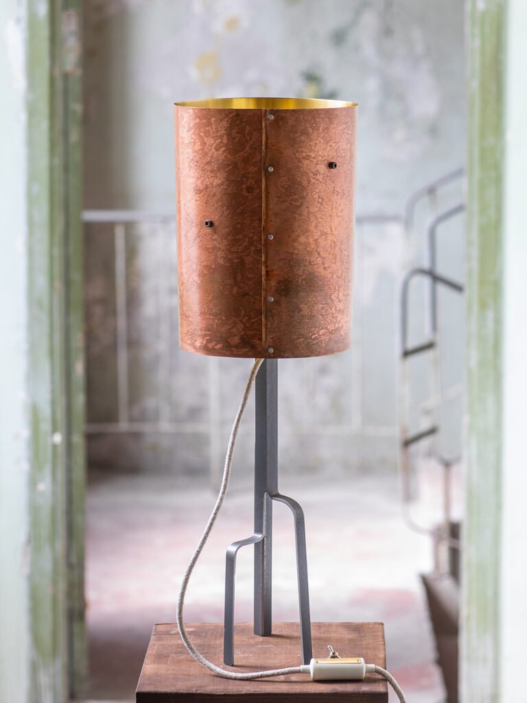 Lamp with a cylindrical brass shade featuring a warm, coppery patina and gold interior, supported by a unique curved three-legged steel stand. Positioned on a wooden surface in a rustic room.