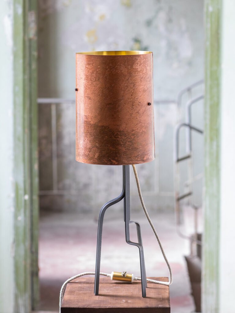 Lamp with a cylindrical brass shade featuring a rich pink patina and gold interior, on a three-legged curved steel stand. On a wooden surface in a rustic room with peeling paint and a green door frame