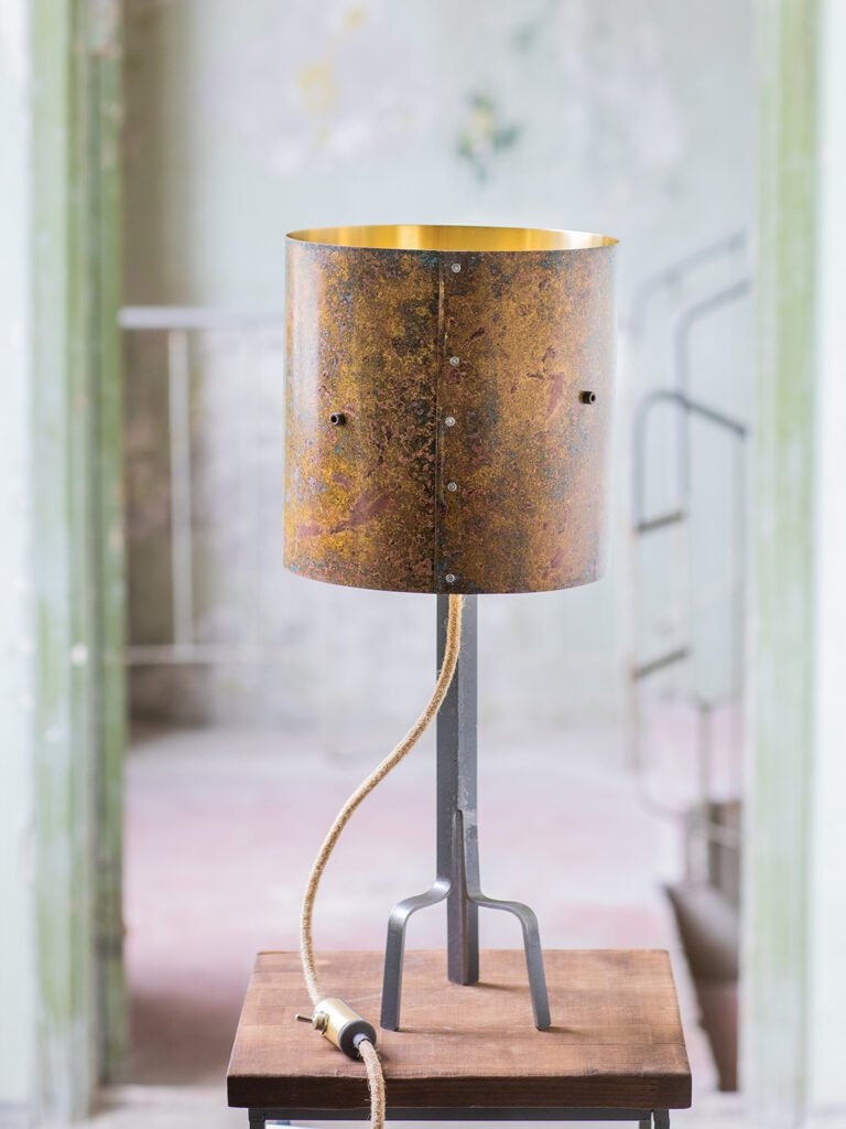 Lamp with a cylindrical, weathered brass shade featuring a textured, rustic finish and gold interior. Mounted on a curved, three-legged steel stand with a visible textile power cord. In a rustic room
