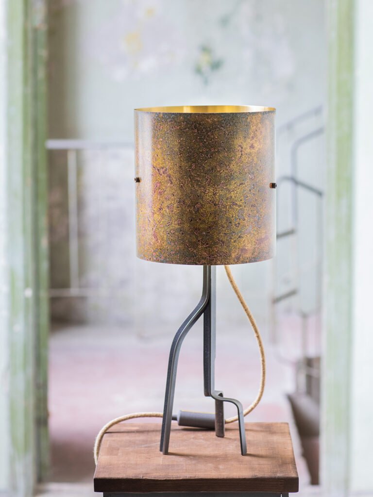 Table lamp with a cylindrical, mottled brass shade featuring earthy tones of brown, green, and gold. Supported by a curved, three-legged steel stand with a textile power cord, set on a wooden surface.