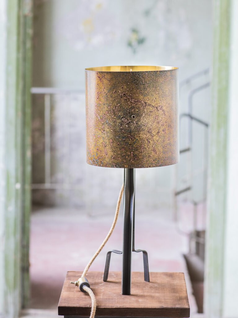 Lamp with a cylindrical, brass shade in earthy tones, featuring a gold interior. On by a three-legged curved steel stand with a beige cord, on a wooden surface in a rustic space with faded walls.