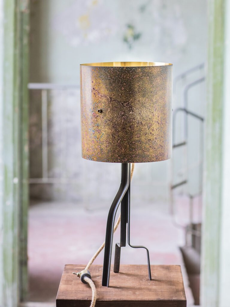 Table lamp with a cylindrical, textured brass shade featuring a rustic patina and gold interior Supported by a three-legged curved steel stand, placed on a wooden surface with a vintage-style backdrop