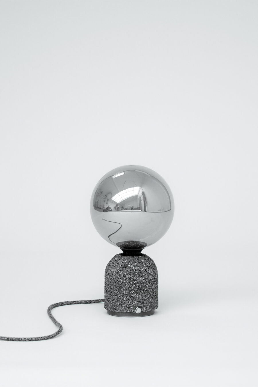 Modern table lamp with a dark aluminum foam base and a mirrored globe bulb, shown against a plain white background with its fabric-covered power cord extending outward.