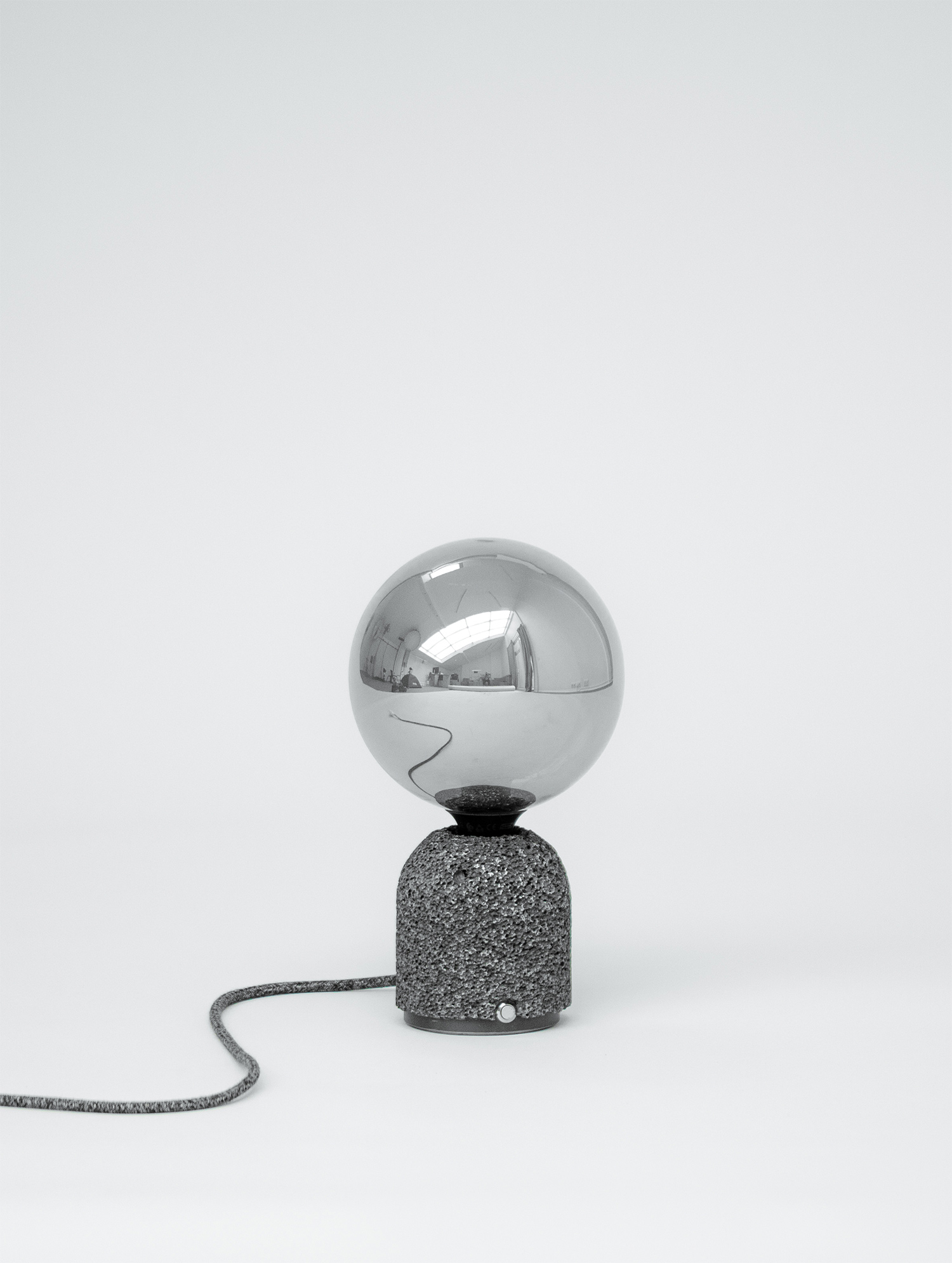 Modern table lamp with a dark aluminum foam base and a mirrored globe bulb, shown against a plain white background with its fabric-covered power cord extending outward.