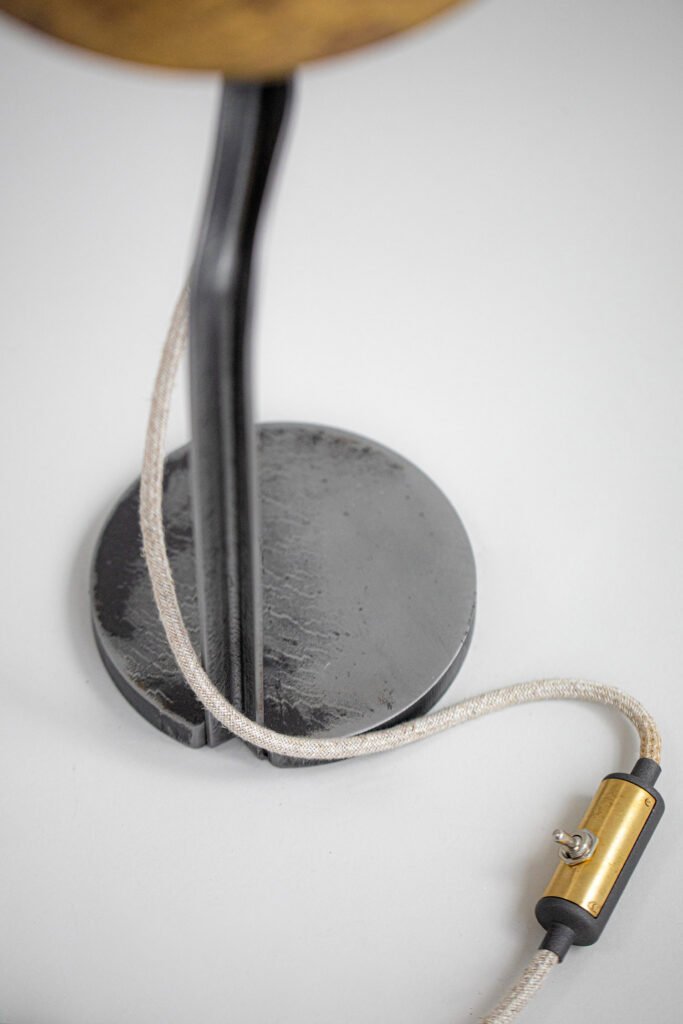 Lamp featuring a black curved steel stand with a round steel base and vintage brass toggle switch on a braided cord. The edge of the patinated brass lampshade is slightly visible at the top