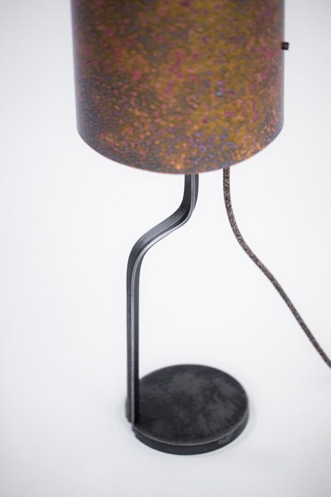 Patinated brass lampshade with speckled textures in brown, purple, and yellow tones, gold interior, angled view showcasing the curved steel stand and base.