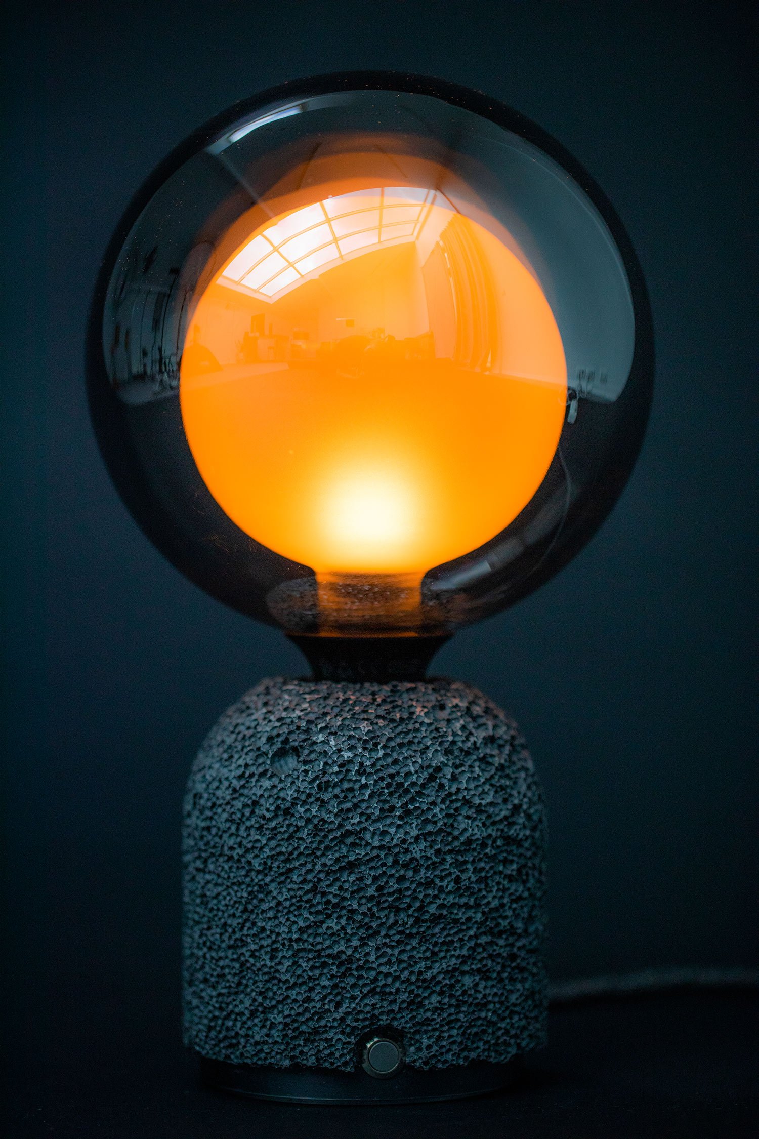 Closeup of a modern table lamp with a dark aluminum foam base and a frosted globe bulb glowing orange from within, set against a dark background