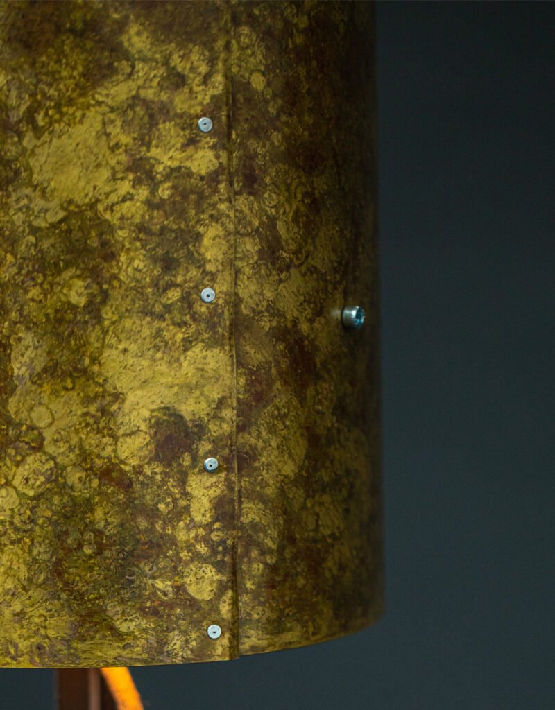 Close-up of a brass cylindrical lampshade with a mottled yellow-brown texture and visible screws securing its design. Set against a dark background, highlighting its weathered appearance.