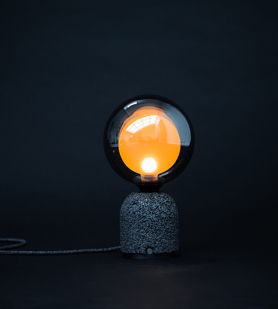 Contemporary table lamp featuring a dark aluminum foam base and a frosted mirrored globe bulb with a warm orange glow from within, set against a dark background