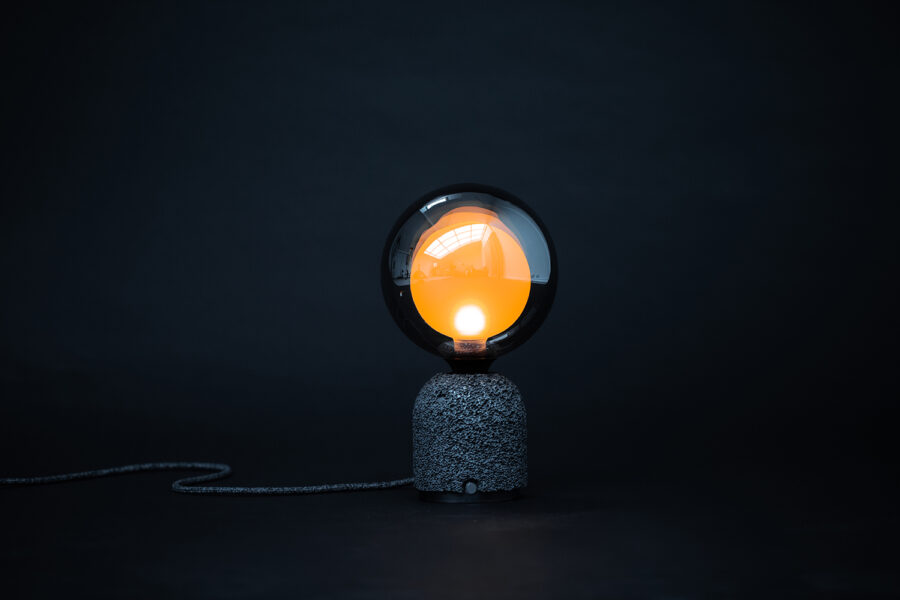 Contemporary table lamp featuring a dark aluminum foam base and a frosted mirrored globe bulb with a warm orange glow from within, set against a dark background