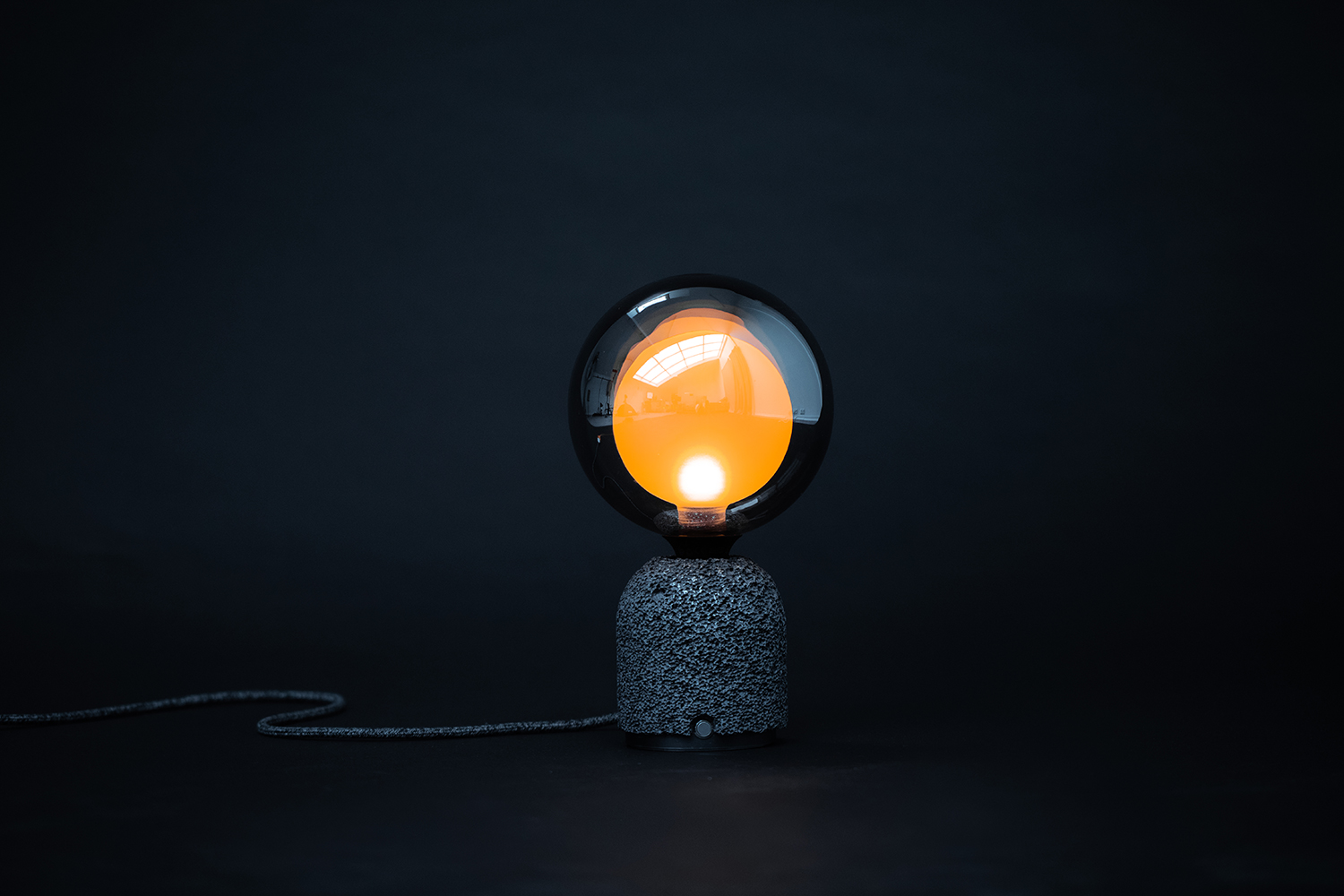 Contemporary table lamp featuring a dark aluminum foam base and a frosted mirrored globe bulb with a warm orange glow from within, set against a dark background