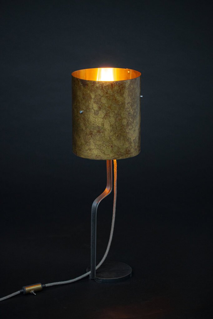 Modern industrial table lamp with cylindrical mottled metal shade emitting warm glow. Curved dark metal stand with round base and fabric cord with visible switch. Set against a dark backdrop.