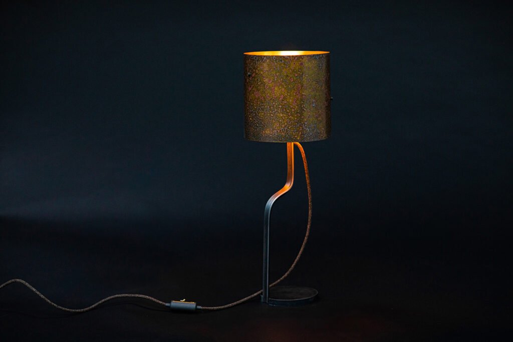 Modern table lamp with a rustic cylindrical shade featuring textured patinated brass in brown and green tones, curved metal stand, and round base. Warm light is emitted from the lamp.