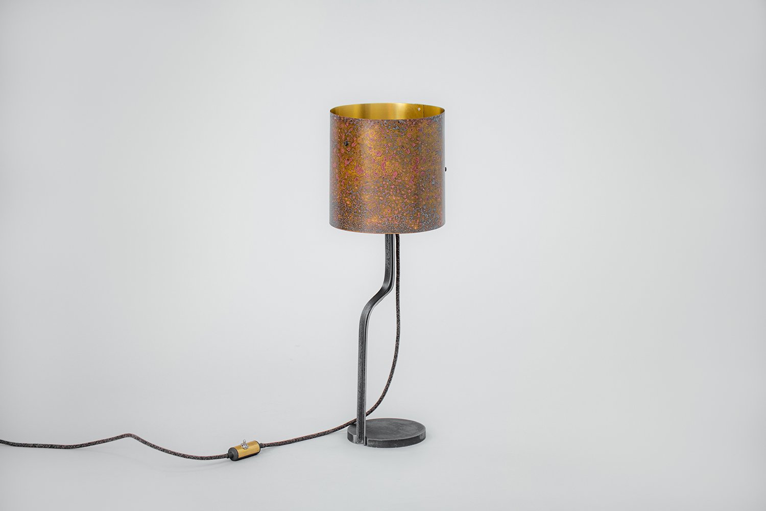 Modern table lamp featuring a patinated brass cylindrical shade in warm hues with a gold interior, curved steel stand, and round steel base