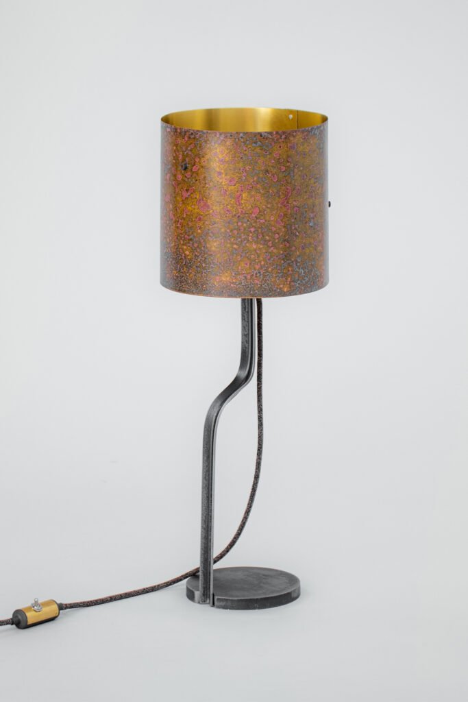 Modern table lamp featuring a patinated brass cylindrical shade in warm hues with a gold interior, curved steel stand, and round steel base