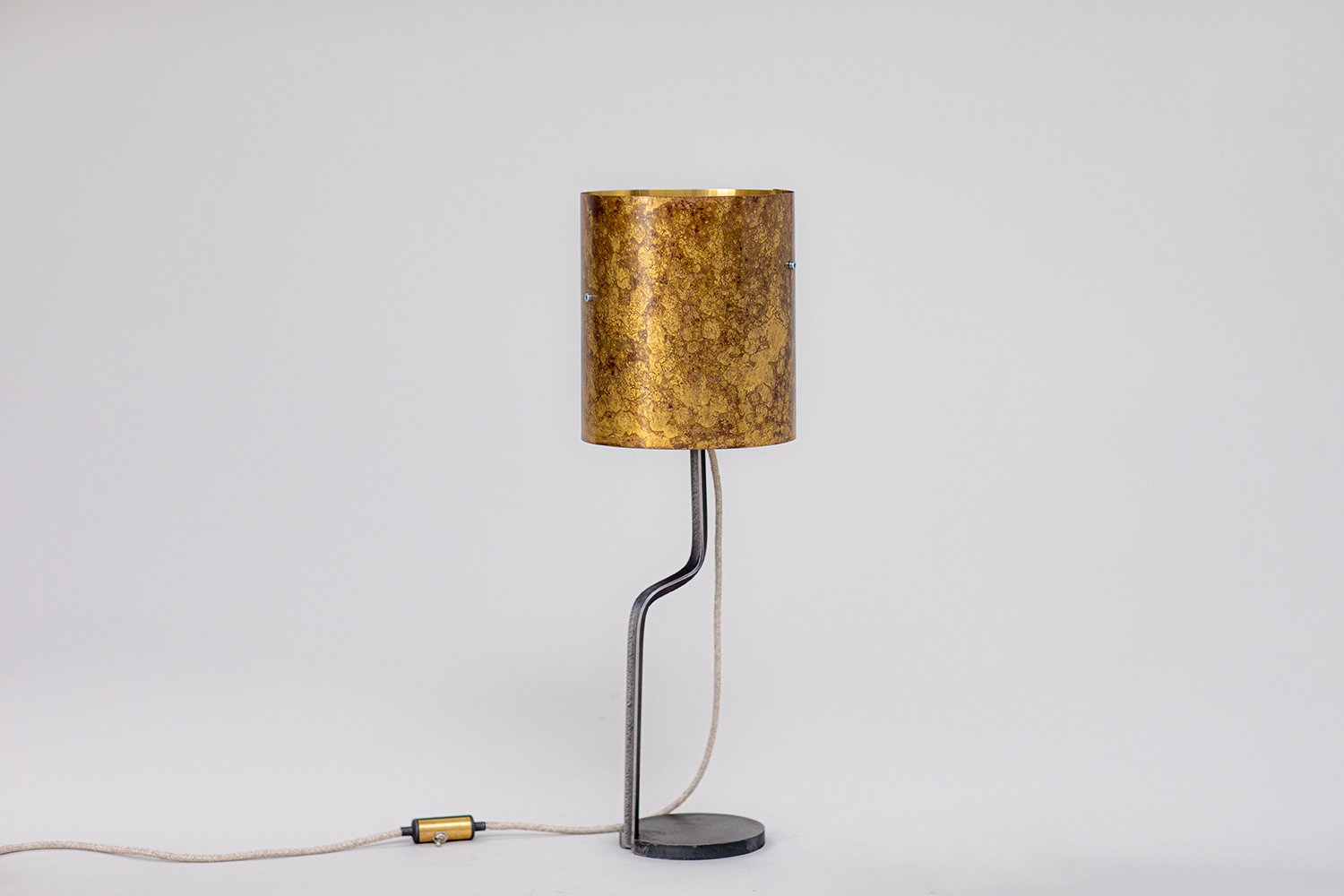 Modern lamp with marbled brass patina cylindrical shade, curved steel stand, round base, and fabric-covered cord with brass inline switch. Presented against a plain white backdrop for emphasis.