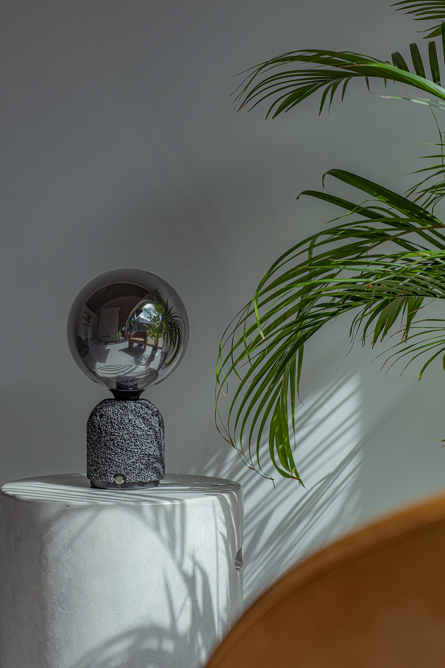 Modern table lamp with a dark aluminum foam base and a mirrored globe bulb, placed on a cylindrical pedestal. A green palm plant casts shadows nearby, and the bulb reflects surrounding furniture and decor in a naturally lit interior