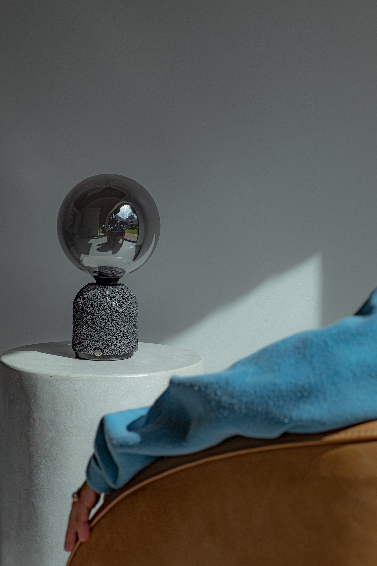 Modern table lamp with a dark aluminum foam base and mirrored globe bulb, placed on a cylindrical pedestal. A seated person in a blue outfit is partially visible in the foreground, with natural light creating soft shadows across the scene