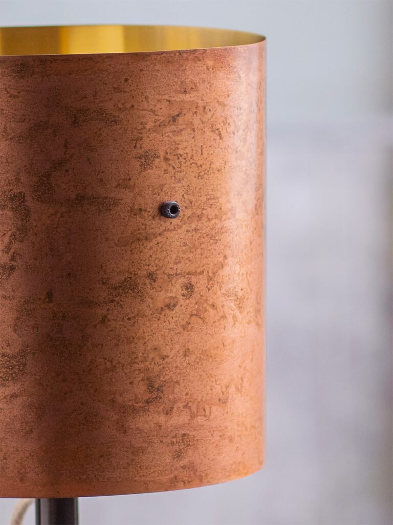 Close-up of a brass lampshade with a textured, reddish-brown surface and a soft blush-pink patina. A black bolt is visible on the exterior, and the interior has a golden hue.