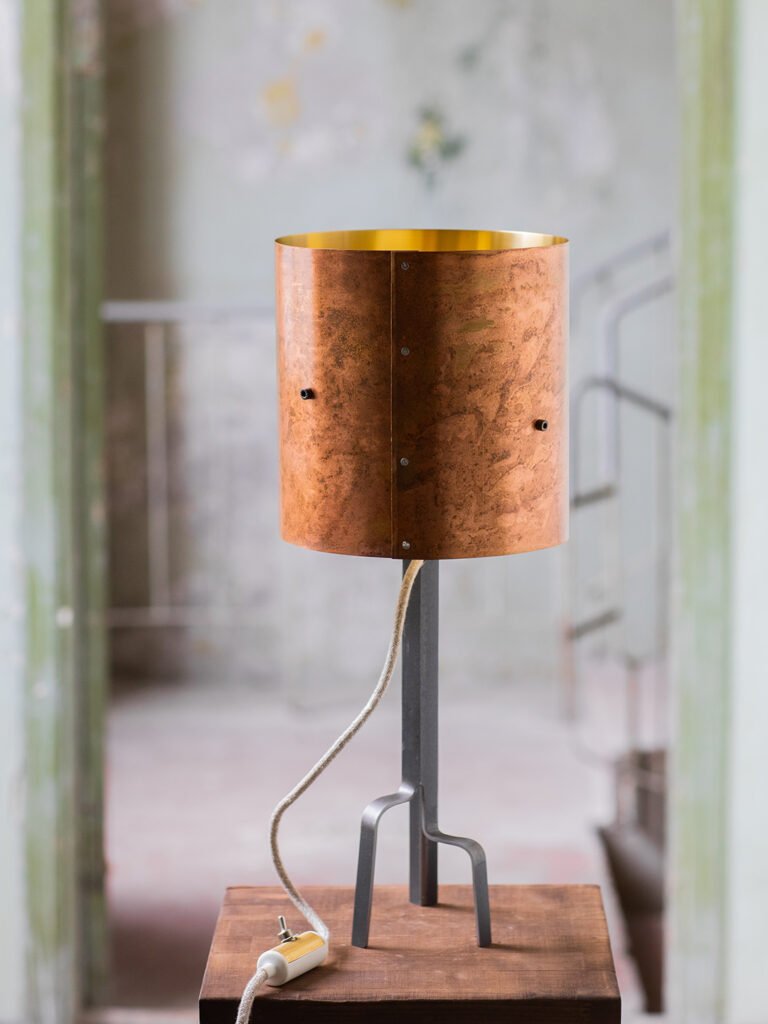 Lamp with a warm rose-gold patina brass shade, gold interior, on a black three-legged curved steel stand. Positioned on a wooden surface.