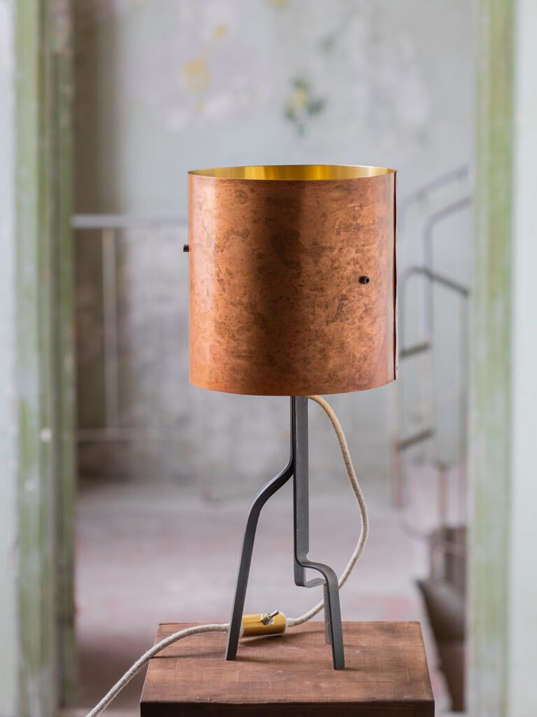 Lamp with a brass shade featuring a warm rose-gold patina and gold interior, supported by a curved three-legged black steel stand. Placed on a wooden surface