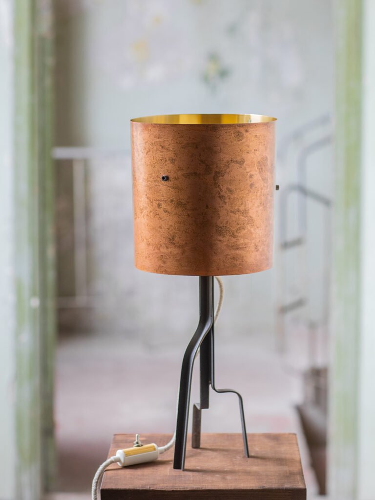 Lamp with cylindrical brass shade with rose-gold patina and gold interior, supported by a curved three-legged black steel stand. Positioned on a wooden surface with a white cord and toggle switch.