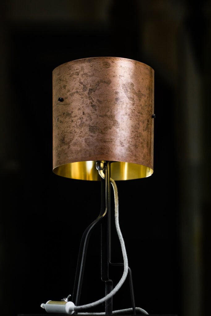 Lamp with a textured, copper-like patinated brass cylindrical shade featuring a golden interior. Mounted on a steel stand, with a visible white braided power cord and toggle switch.