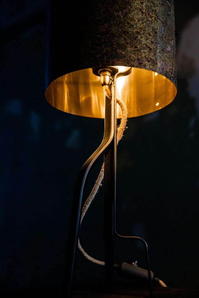 Close-up of a cylindrical brass lampshade with a textured, mottled surface in earthy tones and subtle purple highlights. The glowing golden interior contrasts with the dark background.