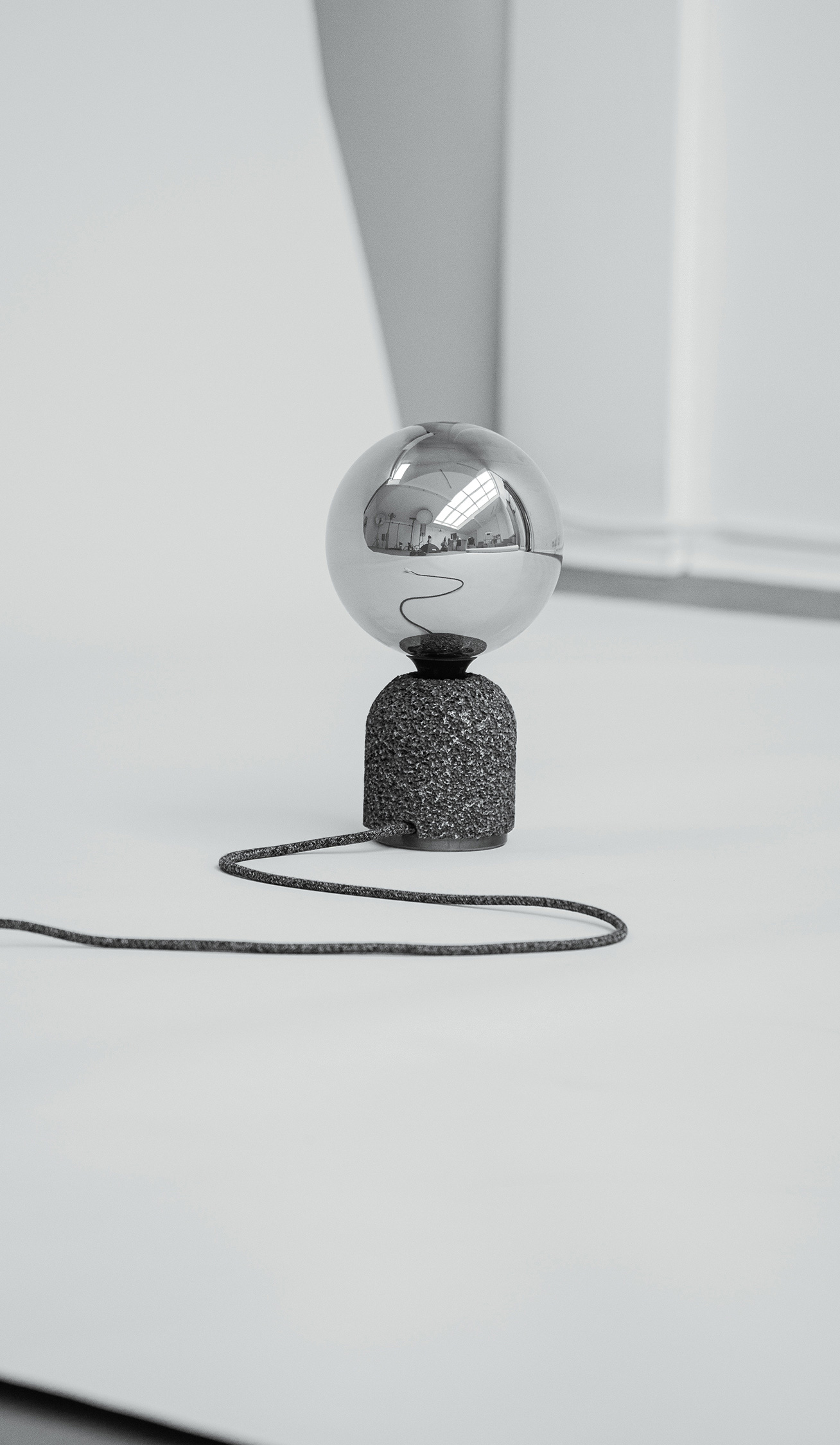 Modern table lamp with a dark aluminum foam base and a mirrored globe bulb, shown in a studio setting. A textile-covered power cable extends from the base and trails across the surface, with windows and equipment reflected in the bulb.