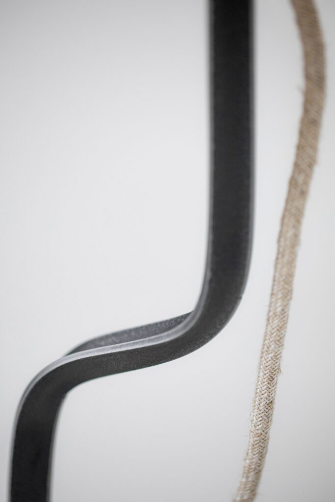 Close-up of a lamp’s dark matte steel stand with a sleek, curved design. A woven textile cable hangs gracefully, showcasing elegant craftsmanship and a balance of durability and style.