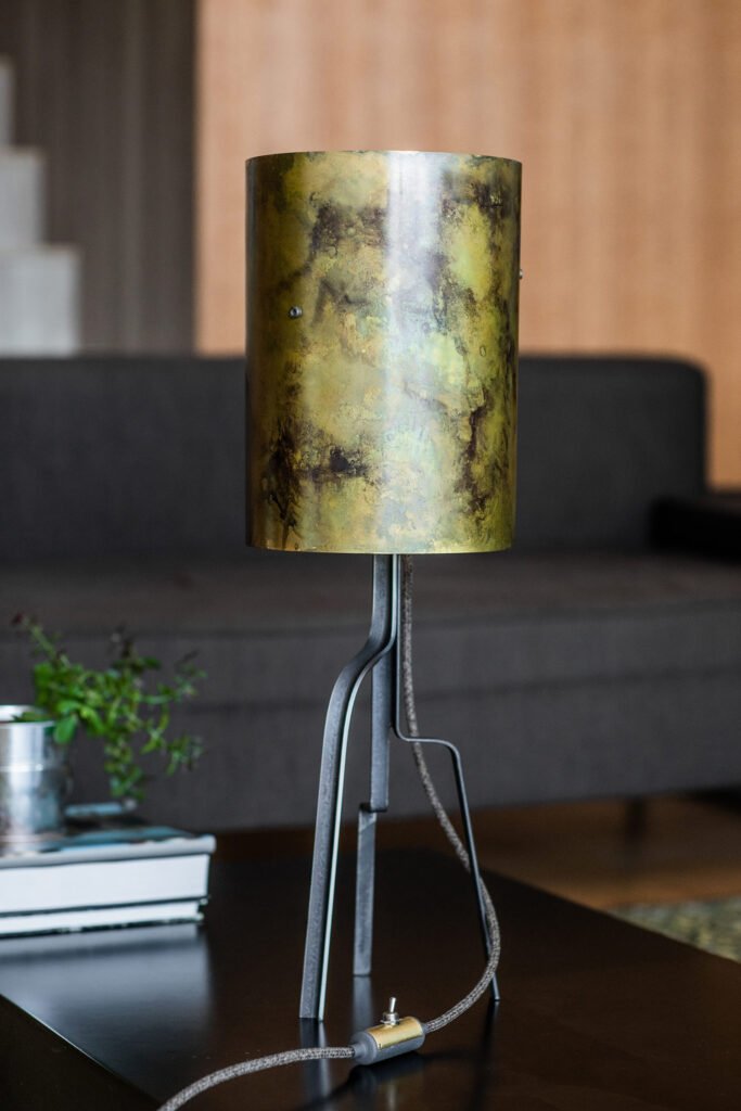 Modern table lamp with cylindrical mottled brass shade in green and gold tones. Minimalist design with slender black frame and fabric cord. Placed on a wooden table with a contemporary backdrop.