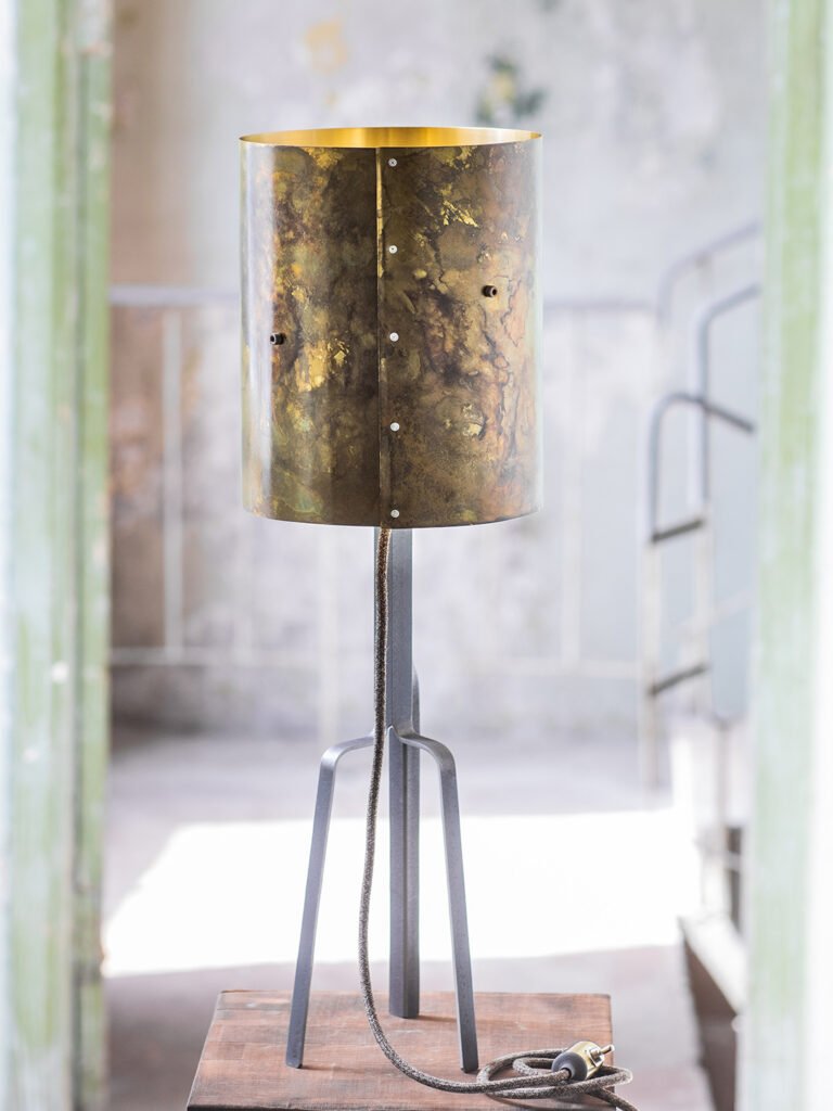 Lamp with textured cylindrical brass shade, mix of gold and brown tones, on sleek tripod stand. Braided cord coiled on wooden surface. Rustic backdrop with faded walls and staircase.