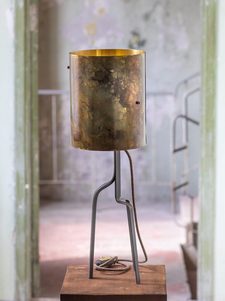 Lamp with cylindrical, textured brass shade on a curved stand. Unlit, with a coiled cord on a wooden surface. Background: rustic interior with worn walls and staircase.