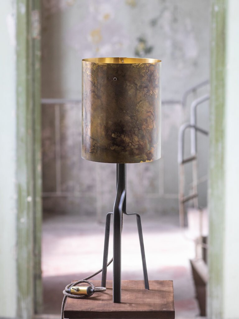 Modern-industrial table lamp with weathered brass shade, grey sculptural base, and brass power switch. Set against aged interior with peeling paint and vintage staircase for an atmospheric look.