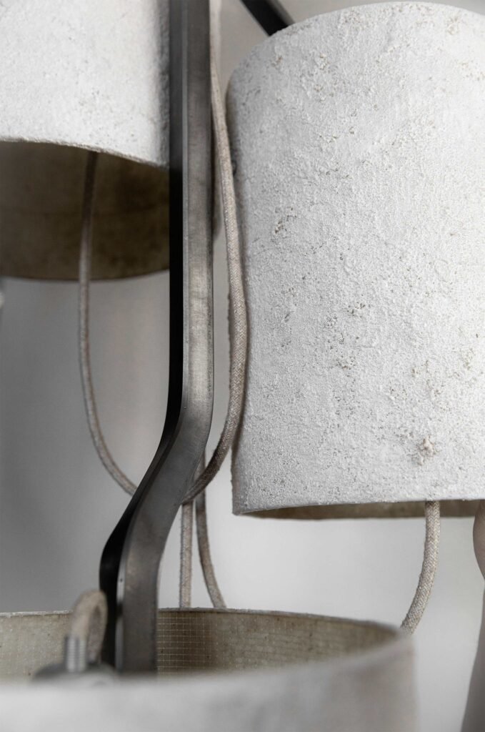 Close-up of a steel chandelier with textured stone-effect cylindrical shades. Textile cables loop around the sleek frame, emphasizing refined craftsmanship and contemporary design.