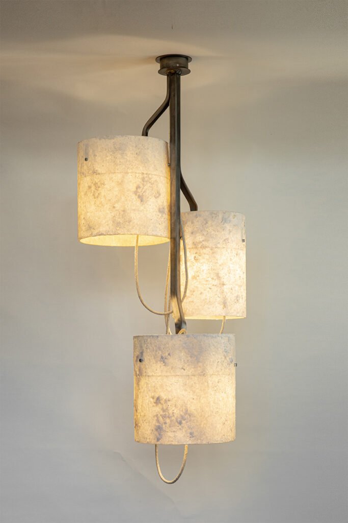 Modern chandelier with three stone-effect cylindrical shades on offset, curved steel branches. Suspended from a circular ceiling rose, it emits soft diffused light, enhancing its elegant design.