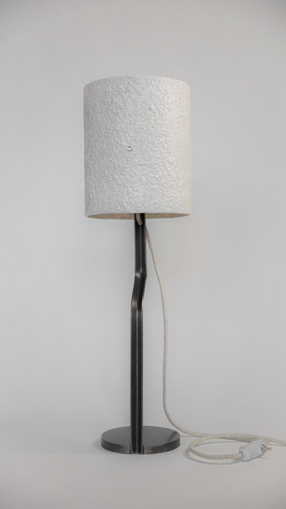 Modern table lamp with a textured white stone-effect shade. Black curved steel stand connects to a round base, with a white textile cord and switch blending style with functionality.