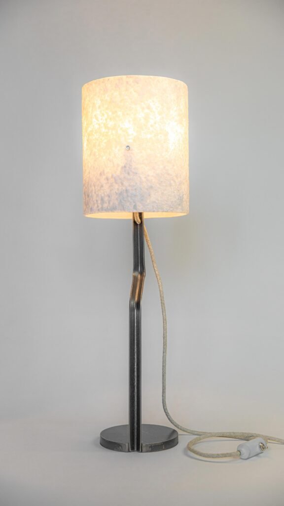 Modern table lamp with a textured white stone-effect shade emitting warm light. Curved steel stand connects to a round base, with a textile cord and switch blending style and function.