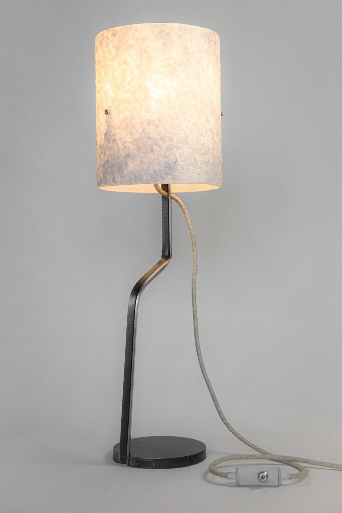 Modern table lamp with a white stone-effect shade diffusing warm light. Angular steel stand connects to a circular base, with a textile cord and inline switch blending style and function.