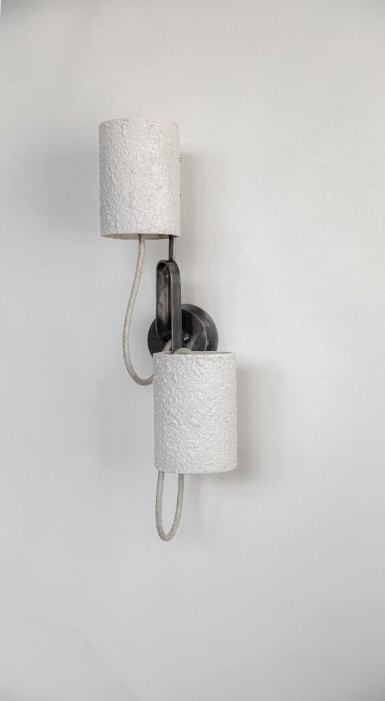 Wall-mounted light with two textured white cylindrical shades on curved metallic arms. A looped cord adds industrial charm, while its modern aesthetic shines as an unlit decorative piece.
