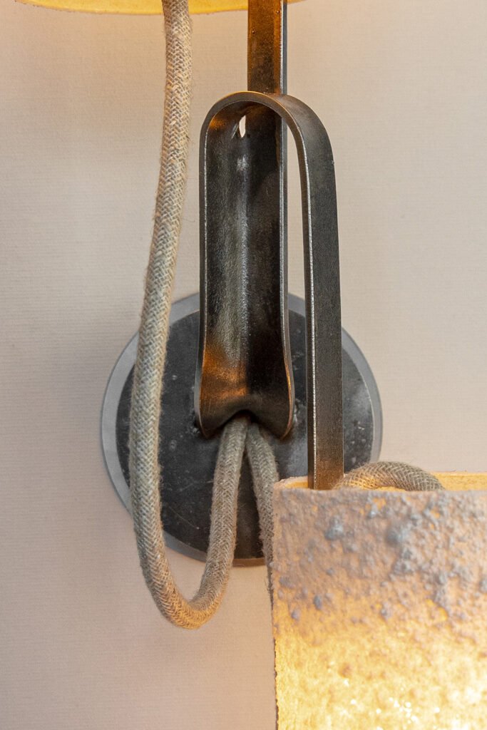 Close-up of a modern wall sconce with a stone-effect cylindrical shade. Curved steel arm and textile cable add industrial flair, emitting a warm glow from a circular wall plate.
