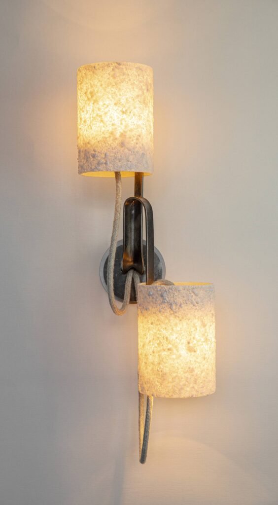 Wall-mounted light with two stone-effect cylindrical shades on curved steel arms. Textile cords enhance its industrial-modern aesthetic, emitting warm diffused light for a cozy ambiance.