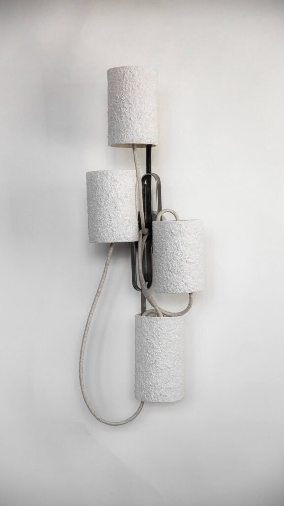 Wall-mounted light fixture with four stone-effect cylindrical shades on curved steel branches extending from a central mount. Textile cables enhance its industrial charm and modern craftsmanship.