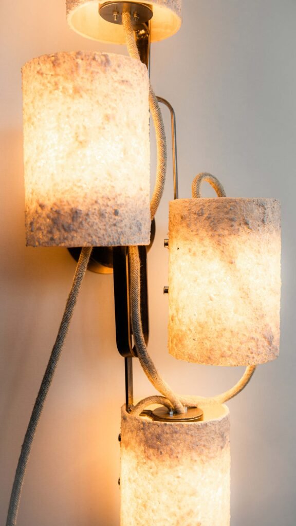 Close-up of a wall-mounted light with three stone-effect cylindrical shades emitting warm light. Curved steel branches and textile cables highlight intricate craftsmanship and industrial style.