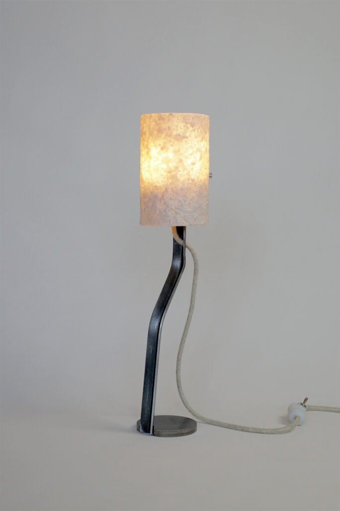 Modern table lamp with a textured stone-effect shade emitting a warm glow. A curved steel stand connects to a circular base, while a textile cord adds practicality to its elegant design.