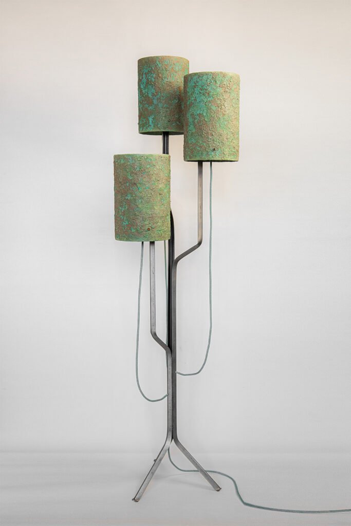 Modern floor lamp with three textured green stone cylindrical shades on a sleek steel frame. Branching arms hold shades at varying heights. Tripod base and neutral wall background complete the design.