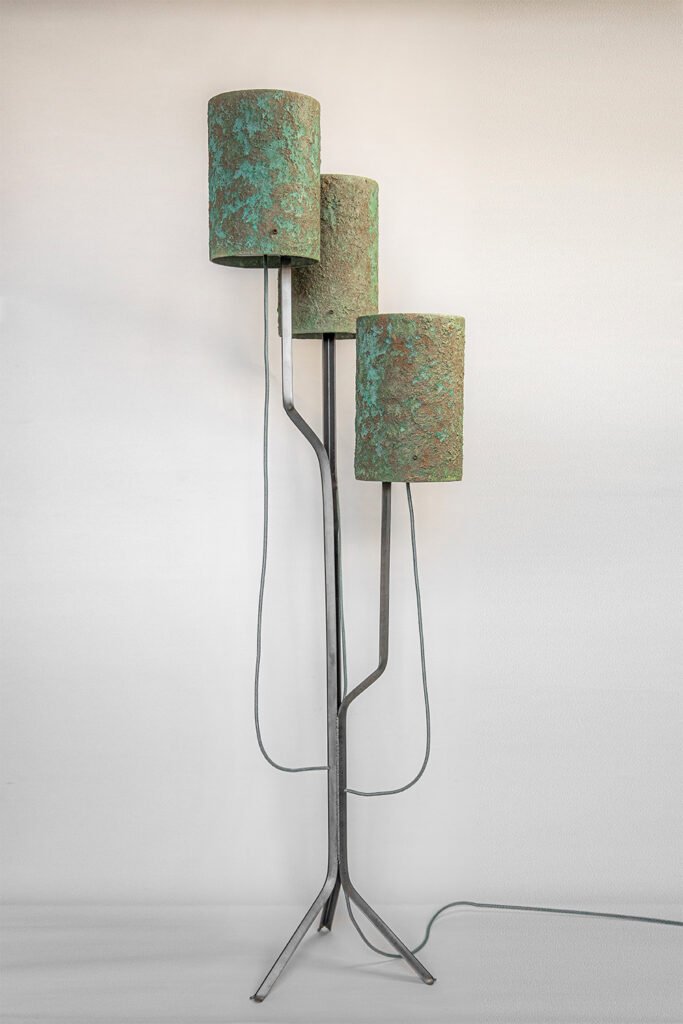 Contemporary floor lamp with three patina-textured cylindrical shades in green and brown. Shades at varied heights on a black angular frame with tripod base. Visible cords and neutral wall background.
