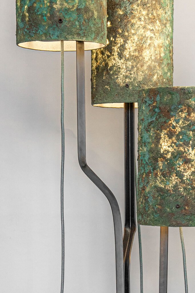 Close-up of a modern floor lamp with three patina-like green and brown textured shades emitting a warm glow. Black angular frame and fabric-covered cord contrast with the neutral wall.