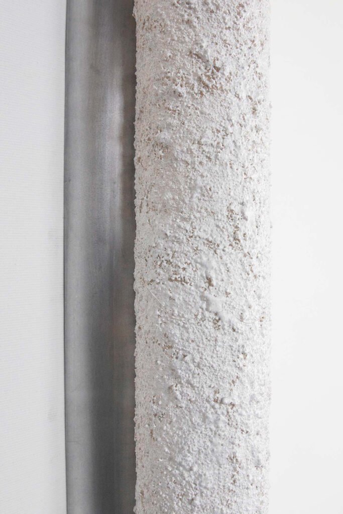 Close-up of a wall-mounted tube light fixture with an unlit textured stone-like surface. Intricate detailing contrasts with the sleek brushed steel rose, blending ruggedness with refined design.